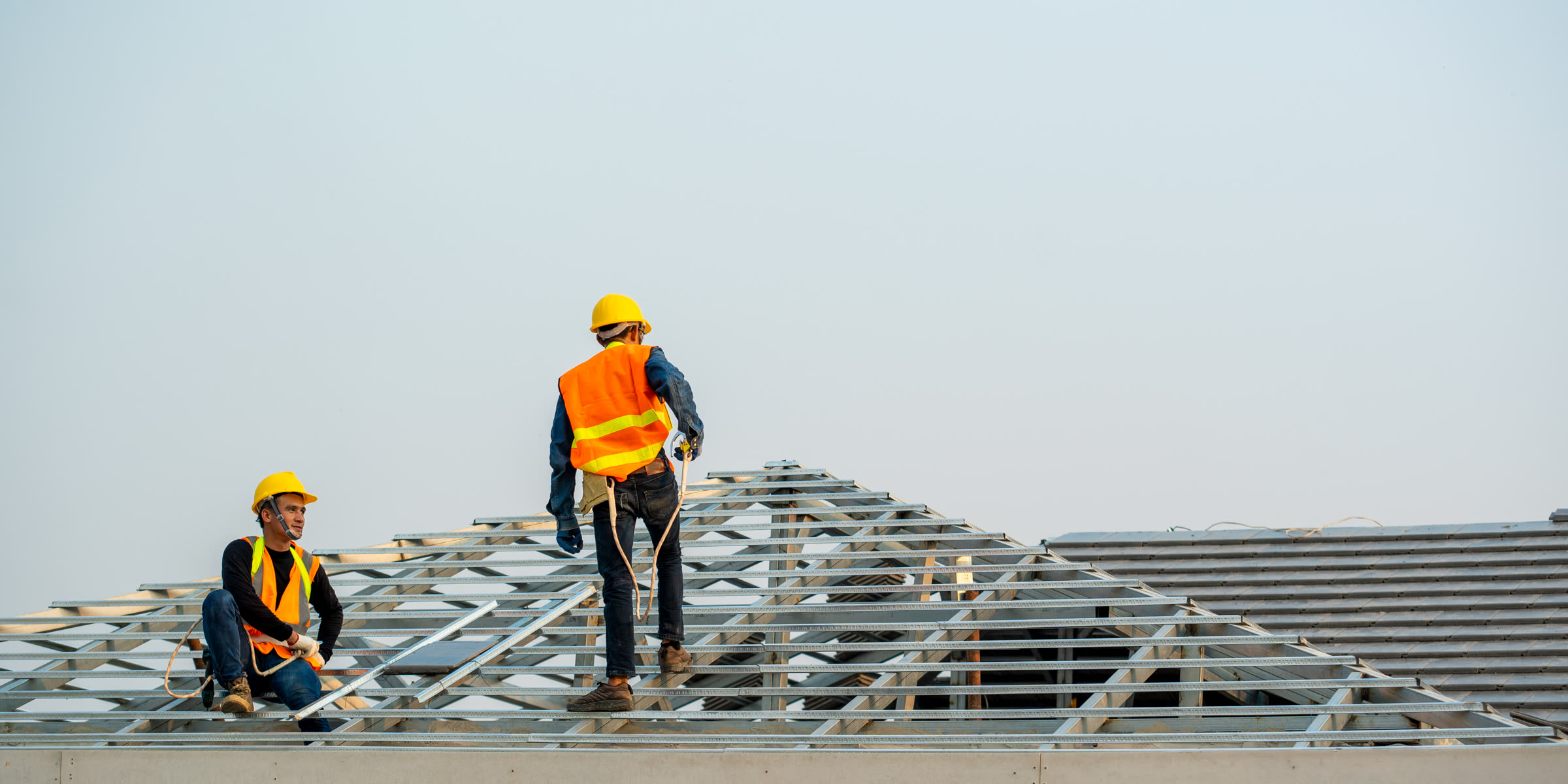 Two roofers in safety vests working on a roof deck against a pale sky
